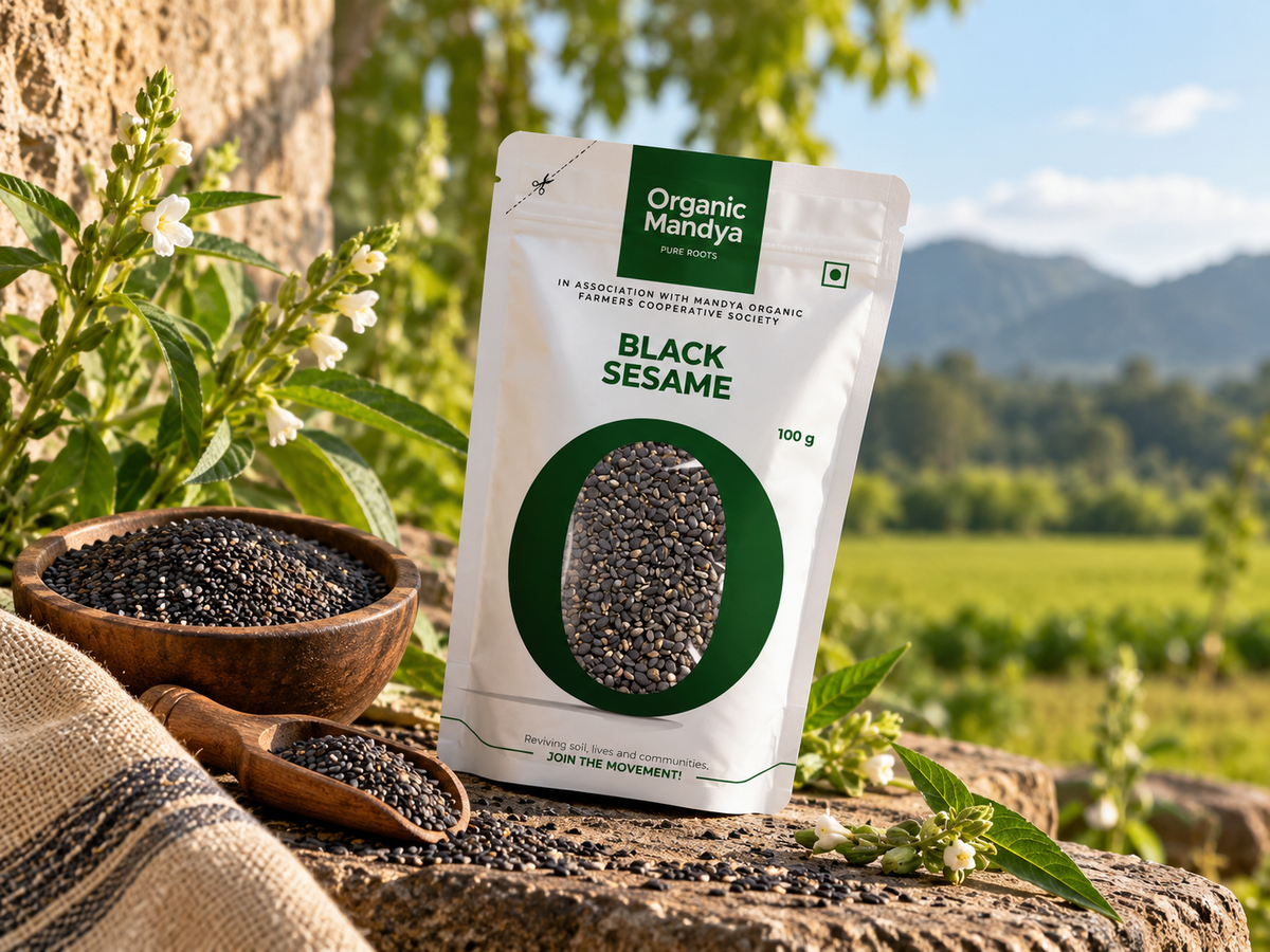 Organic Mandya Black Sesame pouch on a stone ledge beside a wooden bowl and scoop of black sesame seeds, with a flowering sesame plant and green field behind