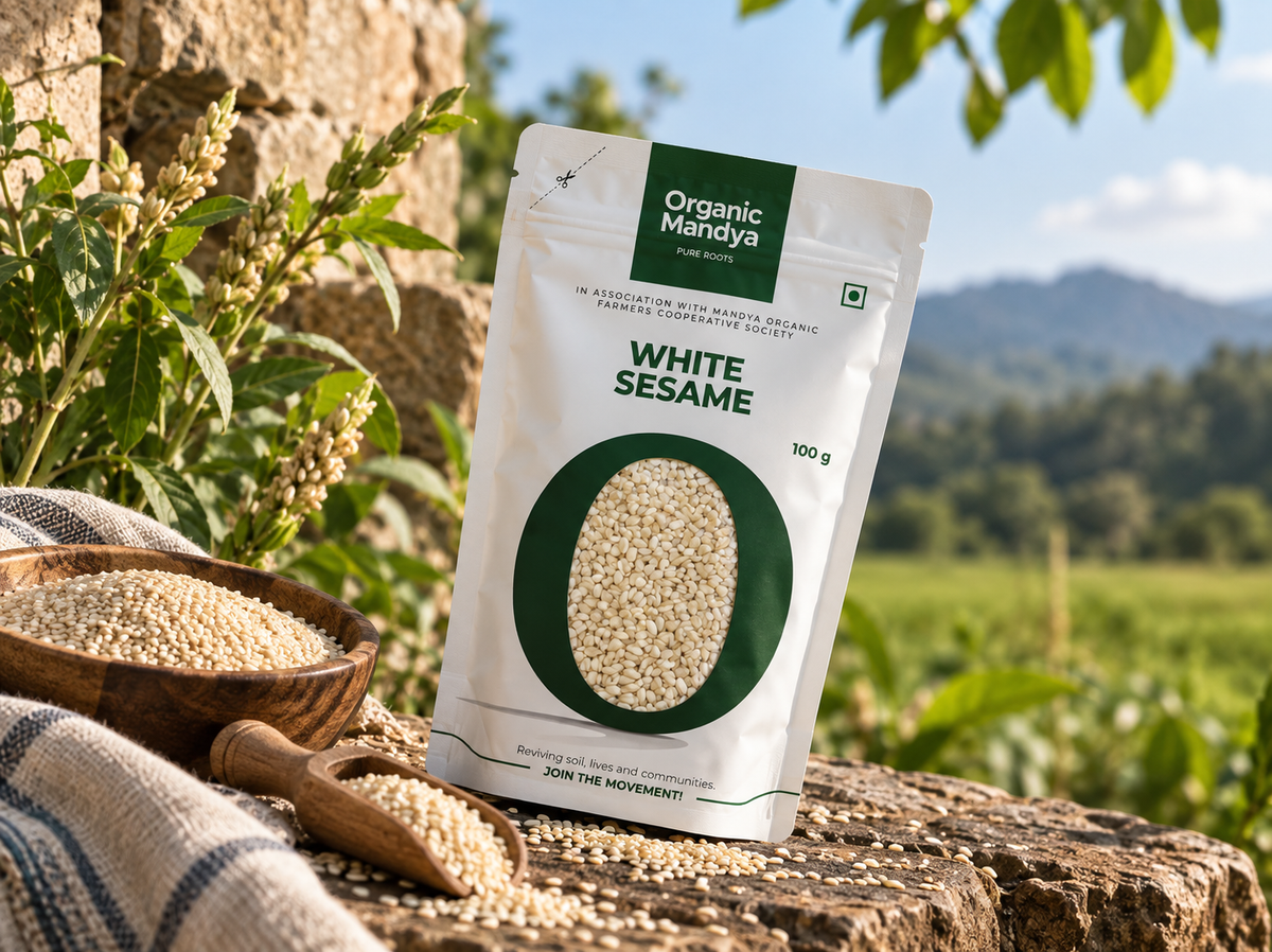 Organic Mandya White Sesame pouch on a stone ledge beside a wooden bowl and scoop of white sesame seeds, with a sesame plant and green field behind